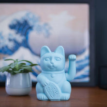 Load image into Gallery viewer, Figurine Maneki Neko chat porte-bonheur bleu clair posée sur une table en bois, devant une œuvre d'art représentant une vague japonaise et une petite plante verte.