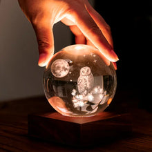 Load image into Gallery viewer, Main tenant délicatement la veilleuse boule de cristal 3D illuminée révélant une chouette et la lune sur son socle en bois de noyer.